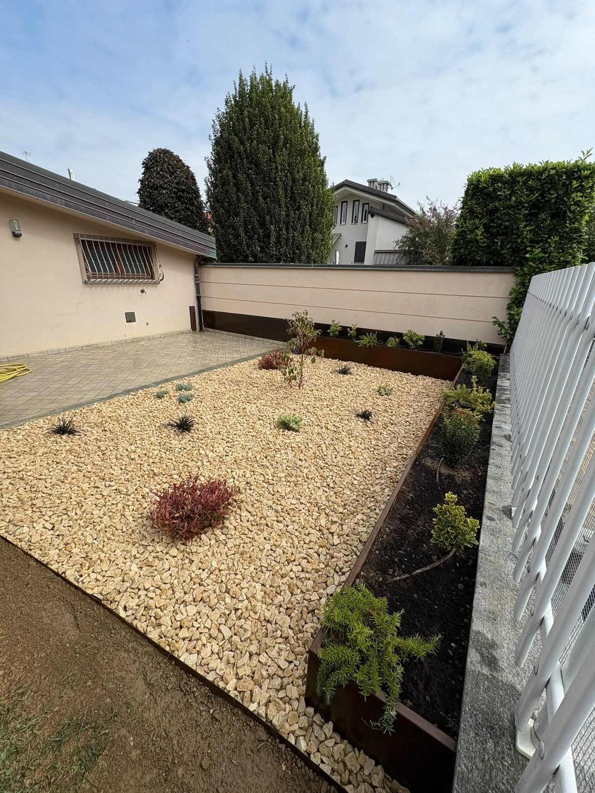 Giardino in ghiaia chiara con piante ornamentali e vasche in corten lungo il muro e la recinzione bianca