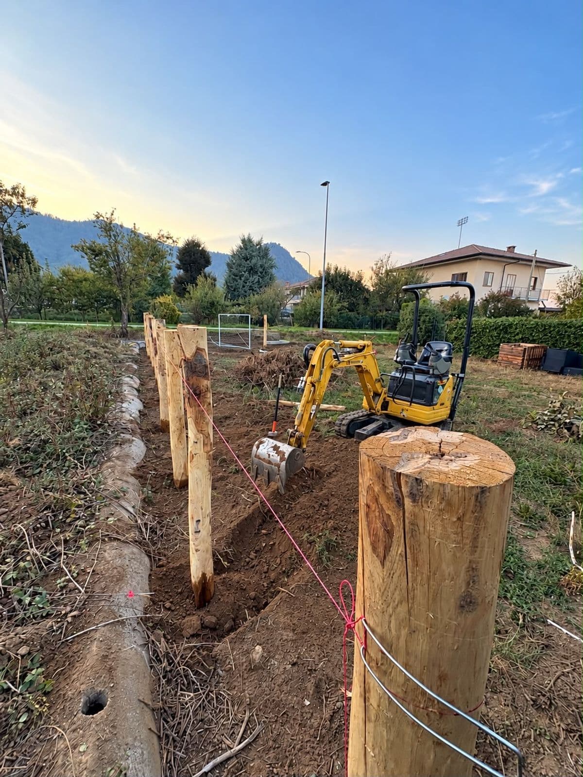 Pali di castagno allineati lungo uno scavo, con il mini escavatore e il filo di tracciamento